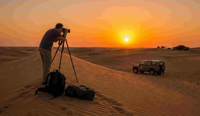 A photographer stands on a sand dune with a tripod and camera, capturing the sunset over the desert landscape with a 4x4 vehicle and camels in the background, illustrating a perfect setting for Arabian desert sunset photography.