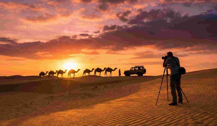 Silhouettes of camels walking across golden sand dunes against a vibrant orange and purple sky, creating a stunning scene for Arabian desert sunset photography.