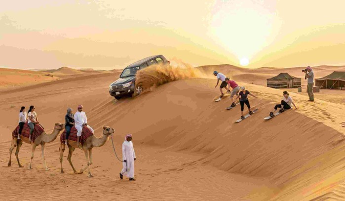 A group of tourists participating in various activities like dune bashing, sandboarding, and camel riding during one of the Best desert safari tours in Jeddah at sunset, with traditional tents in the background.