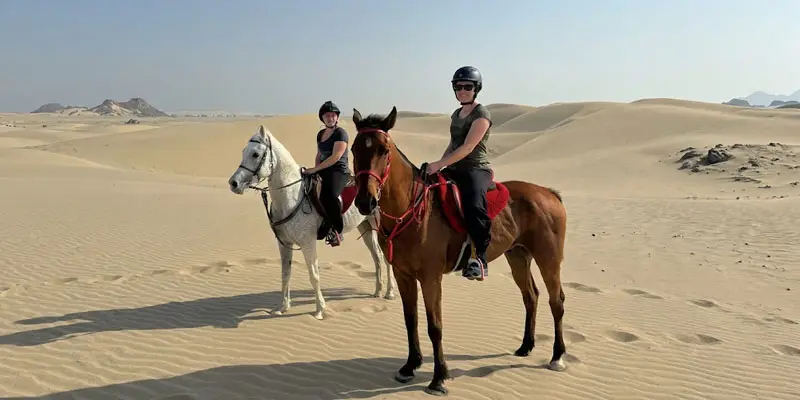 Two female riders wearing helmets posing on horseback atop open sand dunes in the Jeddah desert.