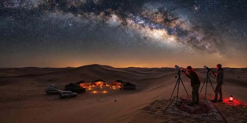 Two astronomers use telescopes on a dune during the Best Time to Go on an Overnight Desert Safari in Jeddah, capturing a stunning view of the Milky Way galaxy and a traditional Bedouin camp below.