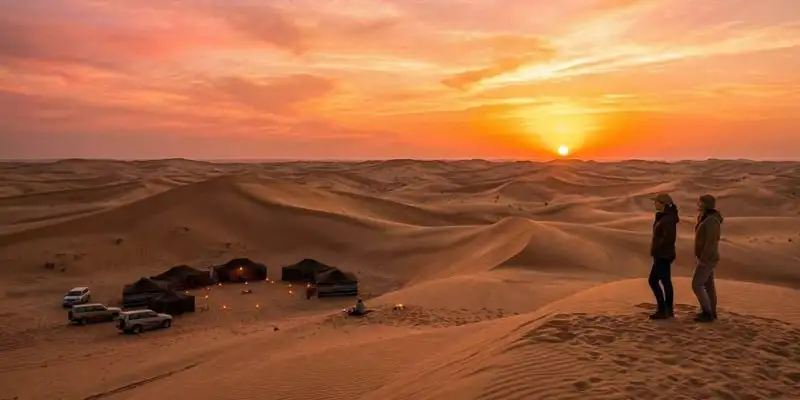 Two travelers stand on a dune at sunset, looking towards a lit traditional Bedouin camp, experiencing the ideal climate during the best time to go on an overnight desert safari in Jeddah.