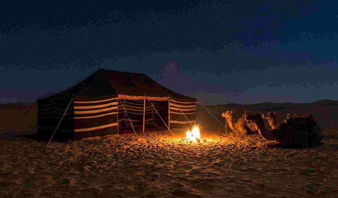 A peaceful desert campsite under a starry sky with Bedouin tents, a campfire, and camels resting nearby, depicting an overnight Camel riding experience Saudi Arabia.
