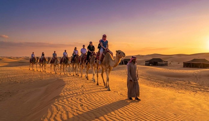 Tourists enjoying a camel ride across the golden desert dunes at sunset, led by a guide in traditional attire, with Bedouin tents in the background, part of an authentic Camel riding experience Saudi Arabia.