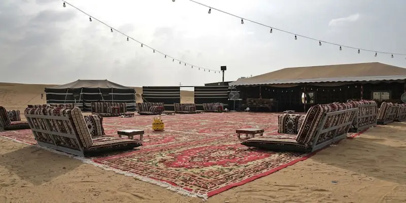 Traditional Bedouin-style desert camp near Jeddah with red carpets, cushioned seating, and overhead fairy lights set up under an overcast sky.