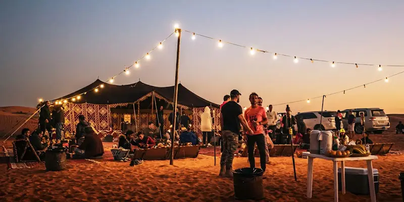 Guests gathering under warm string lights outside a Bedouin tent at an evening desert camp near Jeddah at dusk.