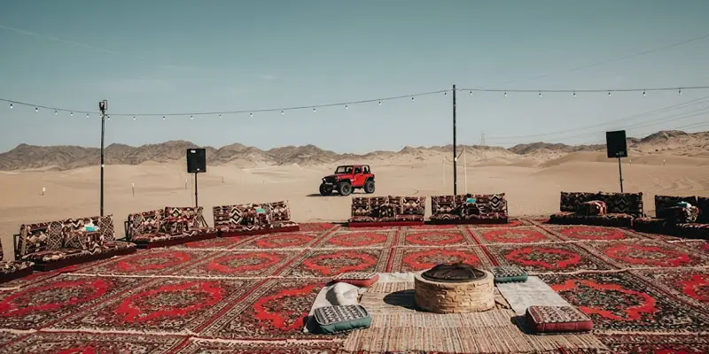 Outdoor desert camp setup in Jeddah featuring a central stone fire pit surrounded by traditional floor seating and a red Jeep Wrangler in the background.
