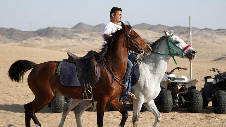 Man riding a brown horse alongside a white horse across the open sand dunes in the Jeddah desert.