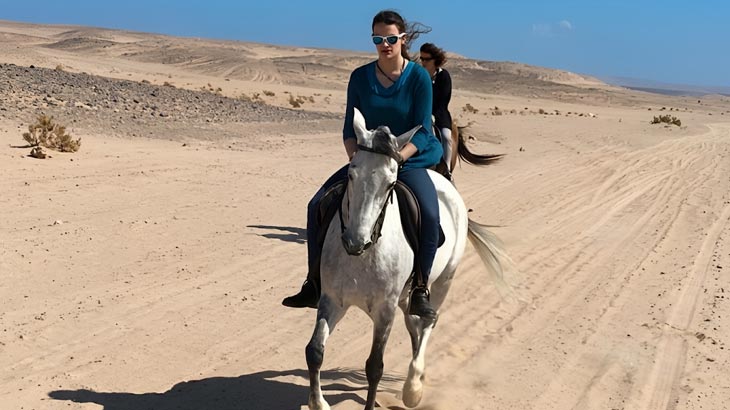 Woman wearing sunglasses riding a white horse along a sandy desert track near Jeddah.