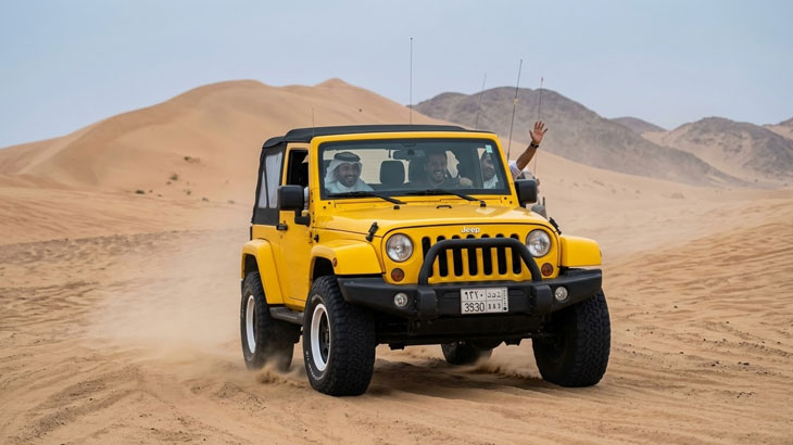A yellow Jeep Wrangler carrying passengers drives through the sandy terrain of the Jeddah desert.