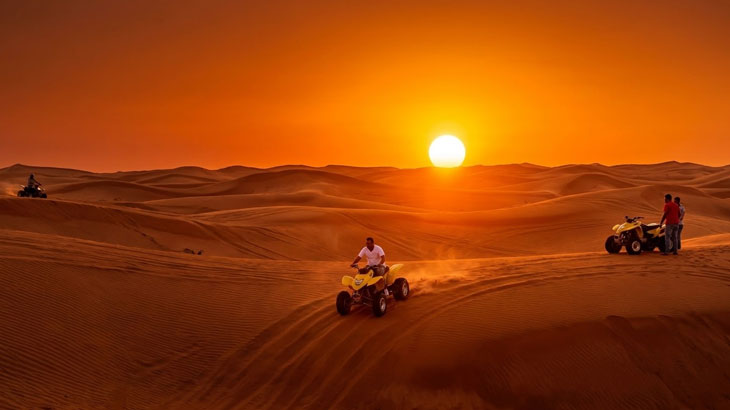 People riding yellow quad bikes over rolling sand dunes during a vibrant orange sunset in the desert.