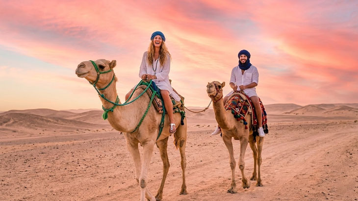 A smiling man and woman riding camels across the Jeddah desert sands under a pink and purple sunset sky.