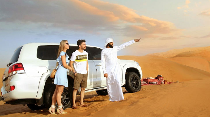 Man in traditional white thobe pointing towards the dunes while a couple stands beside a white SUV in the Jeddah Desert landscape.