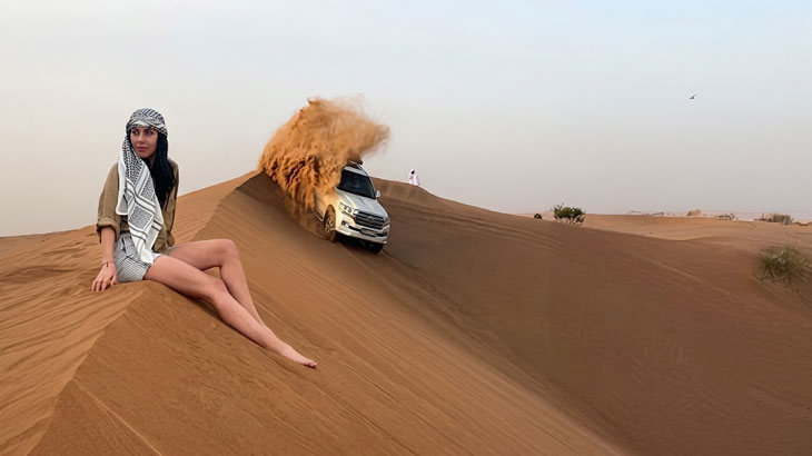 Woman in keffiyeh sitting on a sand dune watching a white SUV dune bashing and kicking up sand in the Jeddah Desert.