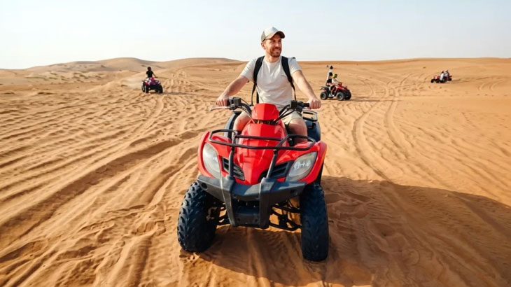 Man smiling on a red quad bike riding towards the camera across vast sand dunes in Jeddah Desert, with other riders in the background.