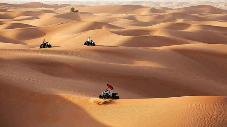 Three quad bikes riding over rolling golden sand dunes in the expansive Jeddah Desert landscape under clear skies.
