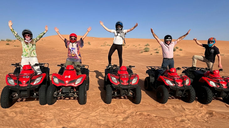 Five happy tourists standing on red quad bikes with arms raised in celebration during a desert tour in Jeddah.